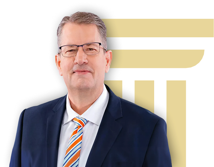 Middle-aged man wearing glasses, navy suit, white shirt, and striped tie against gold pillar design.