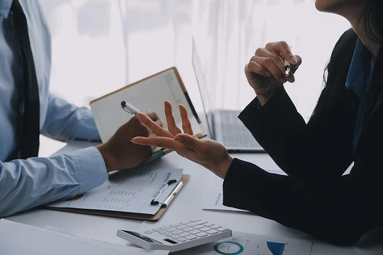 Two professionals engaged in discussion over documents and a clipboard in an office setting.