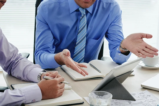 Two professionals in blue shirts discussing documents and a tablet at a desk.