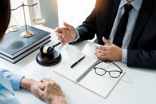 Two professionals in a legal consultation, with gavel, scales of justice, and notebook on table.