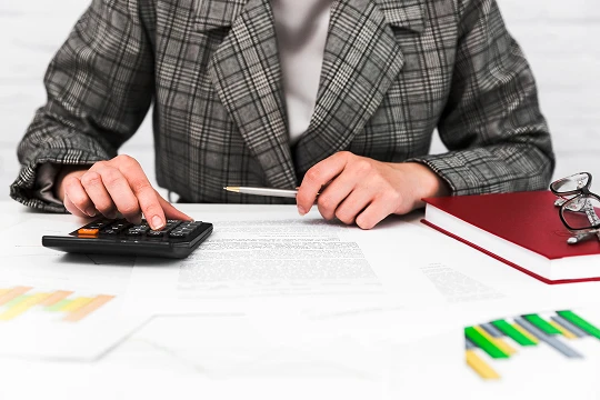 Person in a plaid blazer using a calculator and reviewing documents at a desk.