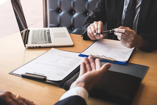 Two professionals in formal attire reviewing documents and a laptop at a wooden table.