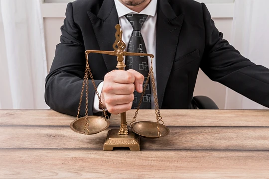 Man in black suit holding bronze balance scale on wooden desk in office.