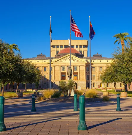 View of a historic government building with three flagpoles displaying the American, state, and POW flags, surrounded by trees and green bollards under a clear blue sky.