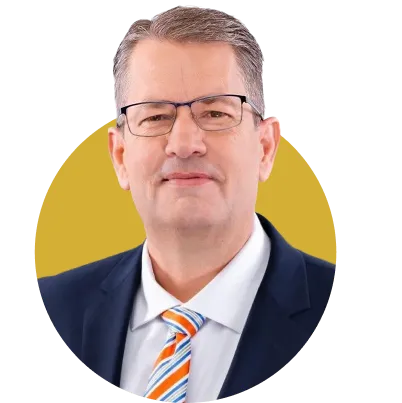 Portrait of a middle-aged man wearing glasses, a navy suit, white shirt, and striped orange and blue tie, set against a mustard yellow oval background.