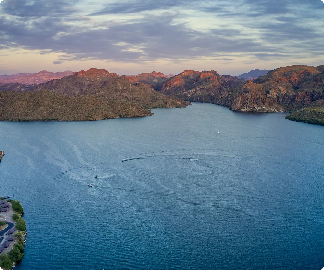 Aerial view of a blue lake with boats and surrounding rocky hills under a cloudy sky at sunset.
