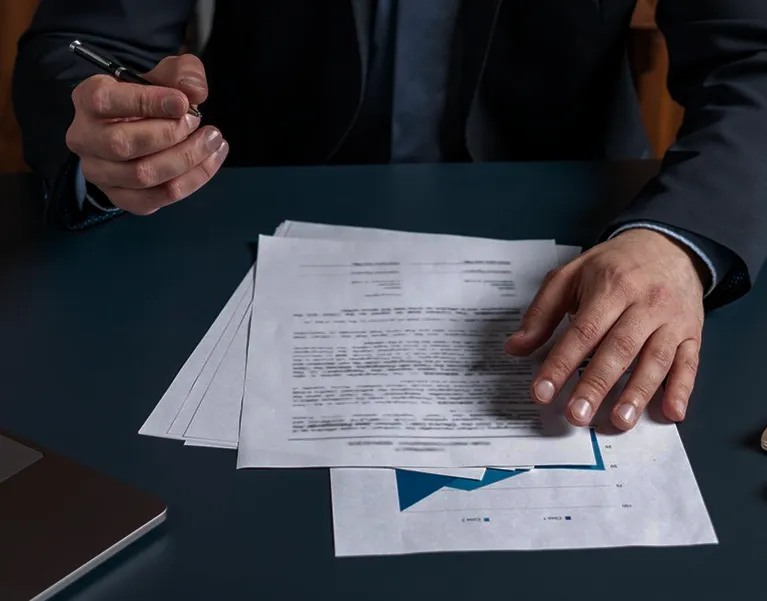 Person in dark suit reviewing and pointing at printed documents on a dark desk.