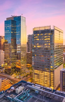 Cityscape at sunset featuring modern glass skyscrapers with illuminated office interiors.