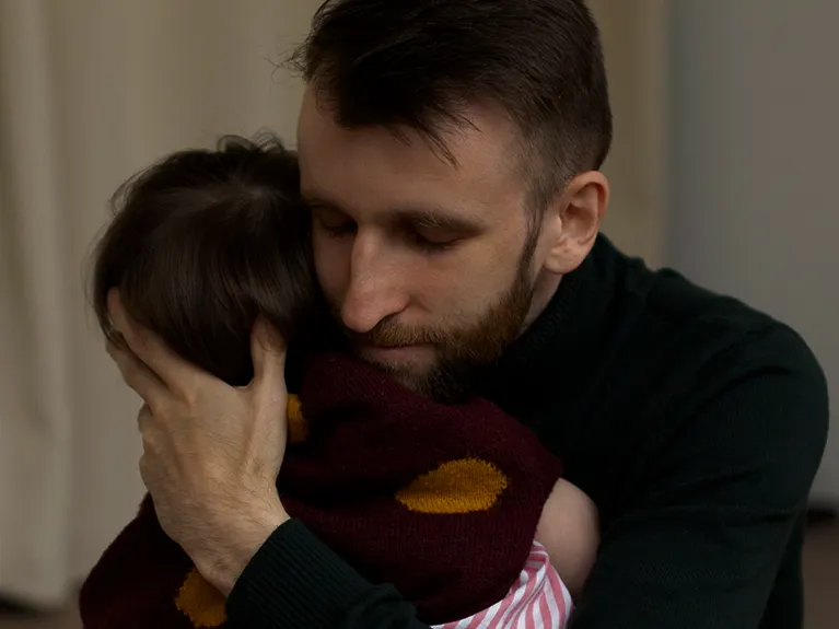 Man with beard tenderly hugging a child wearing a maroon sweater with yellow spots.