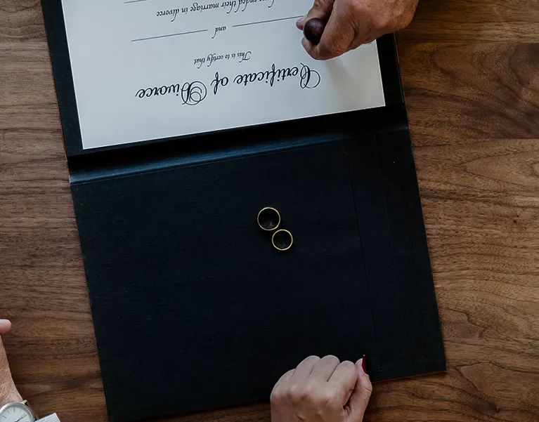 Top-down view of a marriage certificate, two gold rings, and two hands on a wooden table.