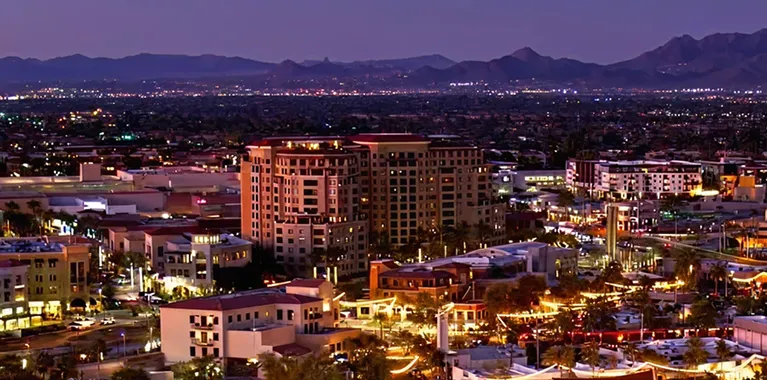 Nighttime cityscape with illuminated buildings, streets, and distant mountains under a purple sky.