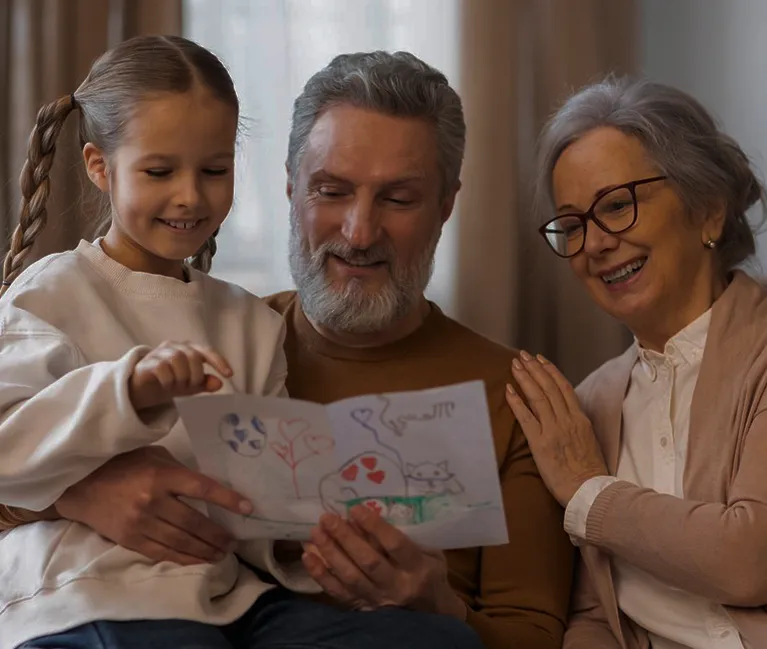 Three generations of a family smiling and looking at a child's colorful drawing indoors.