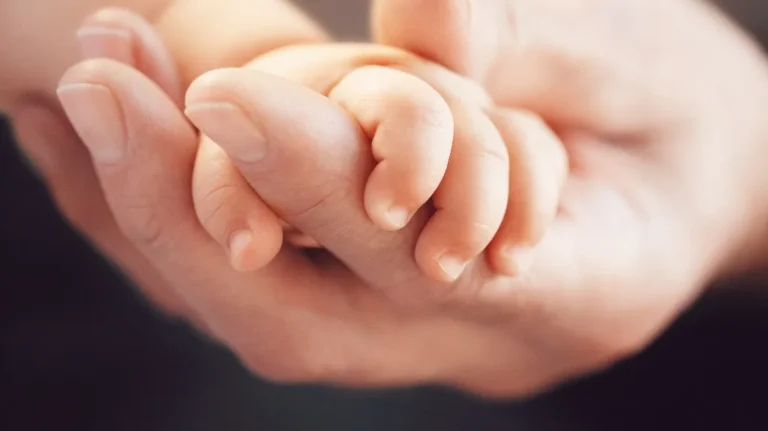 Close-up of an adult hand gently holding a newborn baby's hand against a dark background.