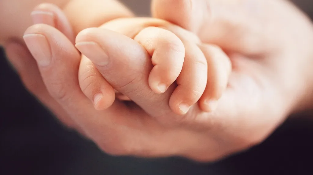 Close-up of an adult hand gently holding a newborn baby's hand against a dark background.