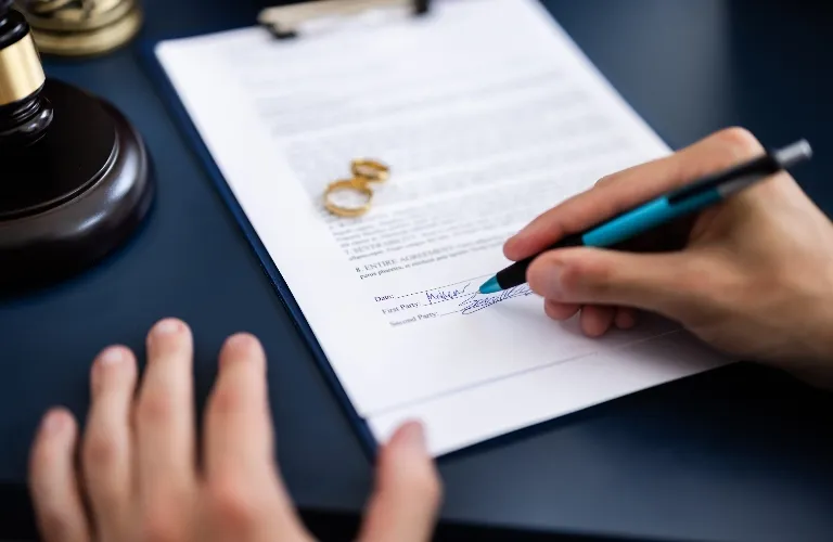 Close-up of a person signing a legal document with two gold wedding rings and a judge's gavel nearby.