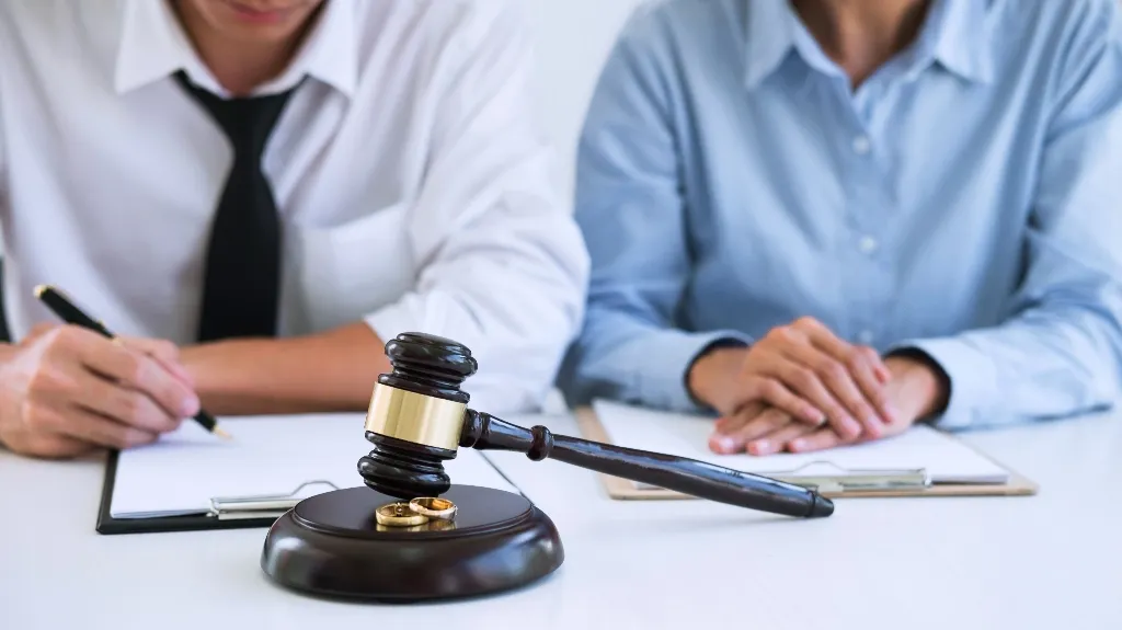 Two individuals in formal attire seated at a table with a judge's gavel and wedding rings in the foreground.