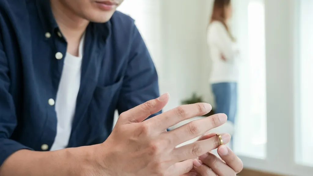 Man in navy shirt removing gold wedding ring, woman blurred in background near window.