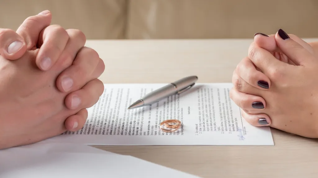 Two pairs of hands clasped over a document with wedding rings and a pen on a light wooden table.