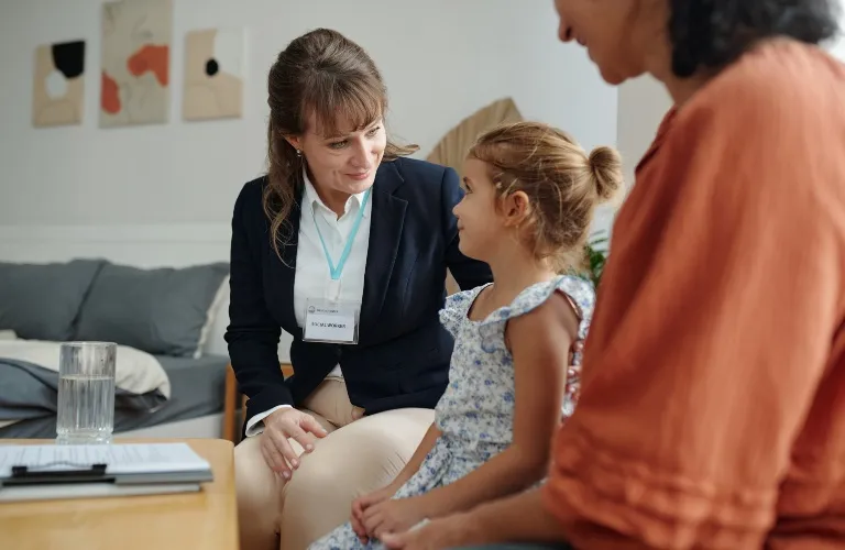 Professional woman with badge speaking gently to young girl, seated beside an adult woman.