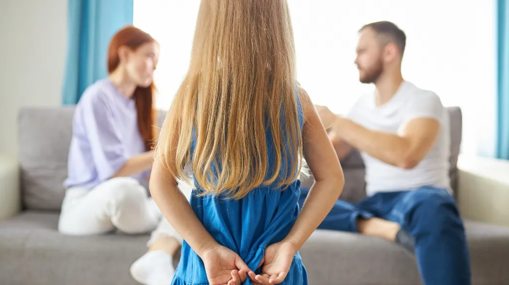 Young girl in blue dress standing with hands behind back, parents arguing on sofa.