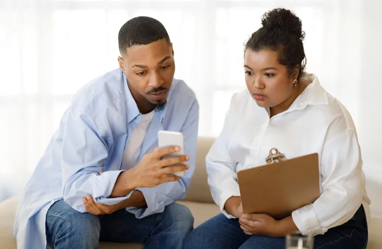Two professionals, a man and a woman, reviewing information on a smartphone and clipboard.