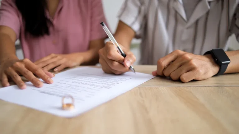 Two individuals seated at a wooden table, one signing a document with a pen, the other pointing at the paper.