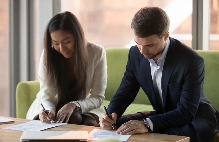 Two professionally dressed individuals, a woman and a man, seated and signing documents on a wooden table.