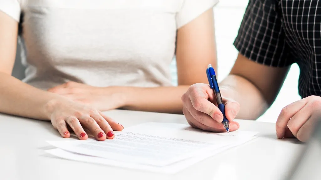 Two individuals at a table, one signing a document with a blue pen, the other observing.