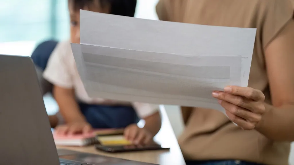 Adult holding and reviewing documents with child using tablet in background.