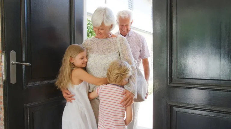 Elderly couple entering home warmly greeted by two young children hugging them.