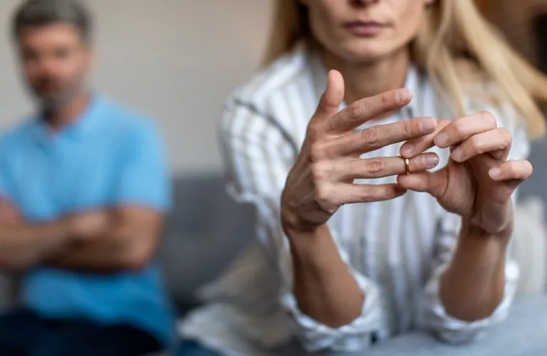 Woman removing wedding ring with blurred man in blue shirt sitting in background.