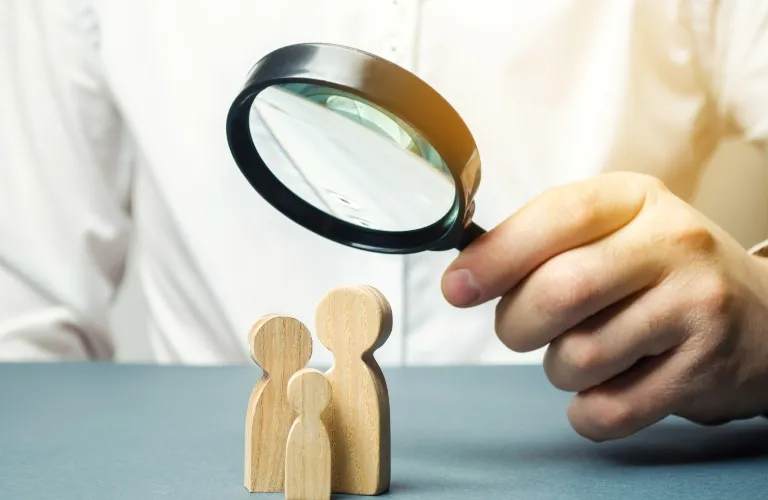 Person holding a magnifying glass over three wooden family figurines on a gray surface.