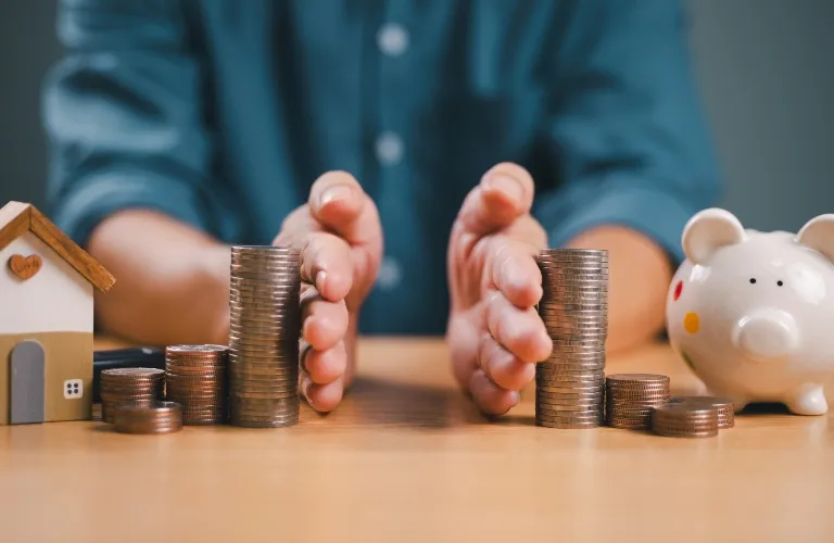 Hands separating stacked coins on a wooden table, with a small house model and a white piggy bank.