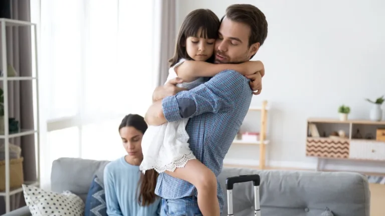 Man in blue plaid shirt holding young girl in white dress, woman sitting on gray couch.