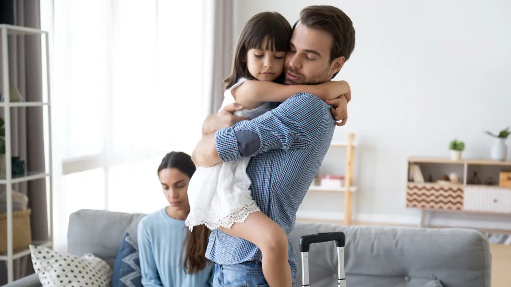 Man in blue plaid shirt holding young girl in white dress, woman sitting on gray couch.