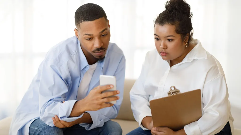 Two professionals, a man in a blue shirt and a woman in a white blouse holding a clipboard, reviewing content on a smartphone.