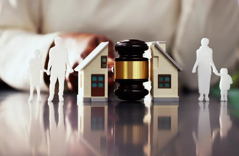 Wooden gavel centered between two miniature houses and white family figurines on reflective surface.