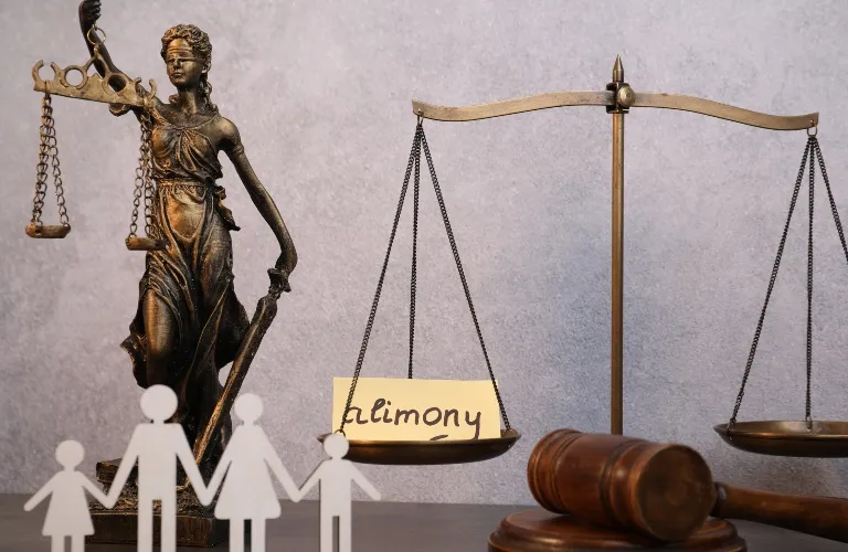 Bronze Lady Justice statue, brass scales with "alimony" note, wooden gavel, and paper family figures on gray background.
