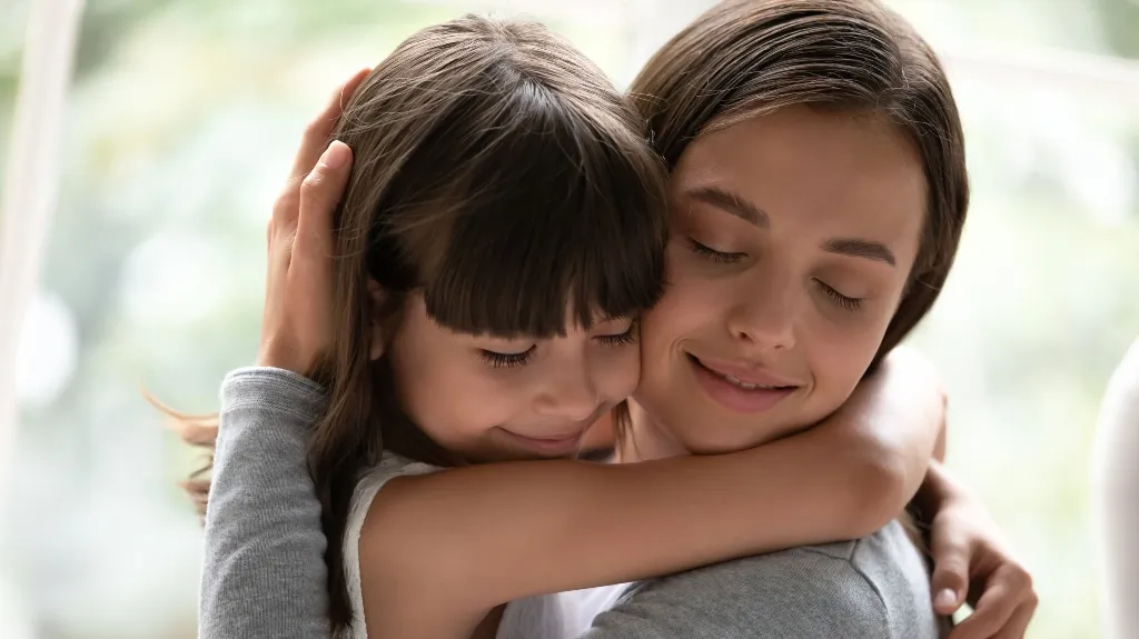 Close-up of a young girl with bangs hugging a smiling woman with closed eyes.