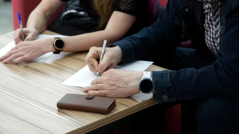 Two individuals writing on white paper at a wooden table, both wearing smartwatches.