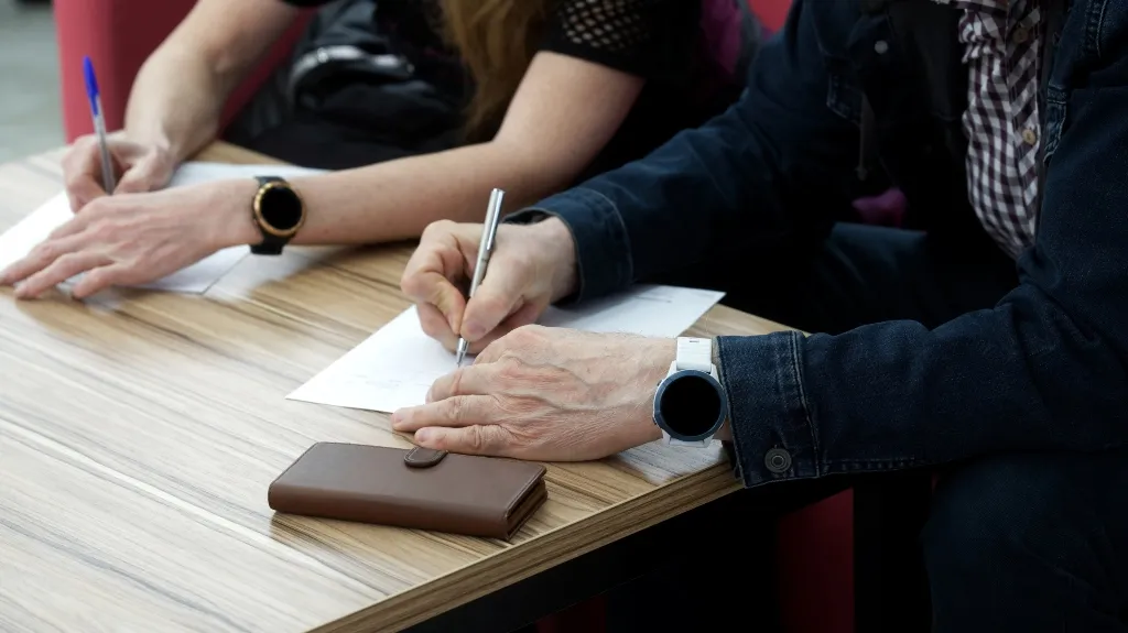 Two individuals writing on white paper at a wooden table, both wearing smartwatches.