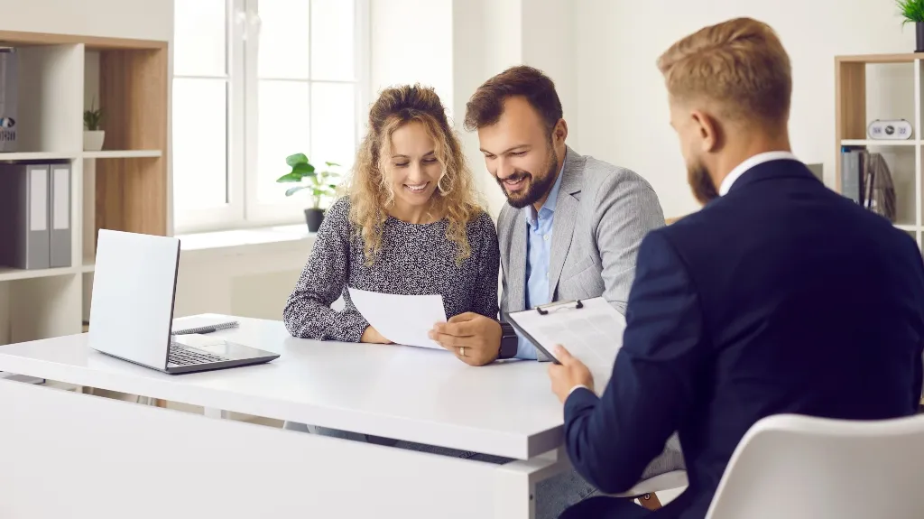 Three business professionals in an office reviewing documents at a white desk with a laptop.
