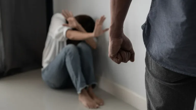 Person sitting on floor shielding face with hands, another person standing with clenched fist.