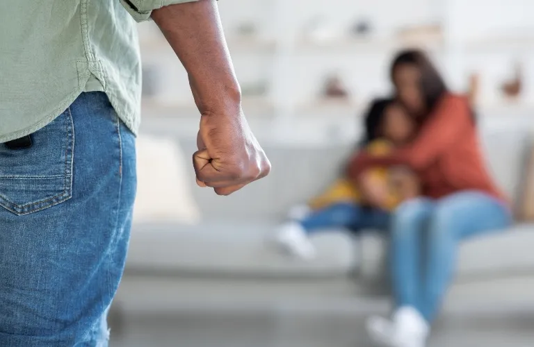 Close-up of a clenched fist in the foreground with a blurred woman and child seated on a sofa in the background.