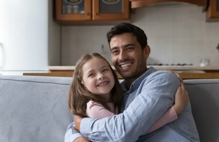 Smiling man in blue shirt hugging young girl with brown hair on gray couch.