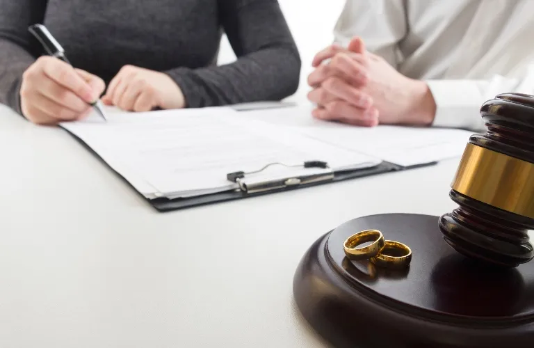 Two people signing documents at a table with a judge's gavel and two gold wedding rings in the foreground.